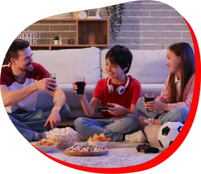 A family of three, sitting on the floor at home, enjoying snacks and drinks while laughing together.