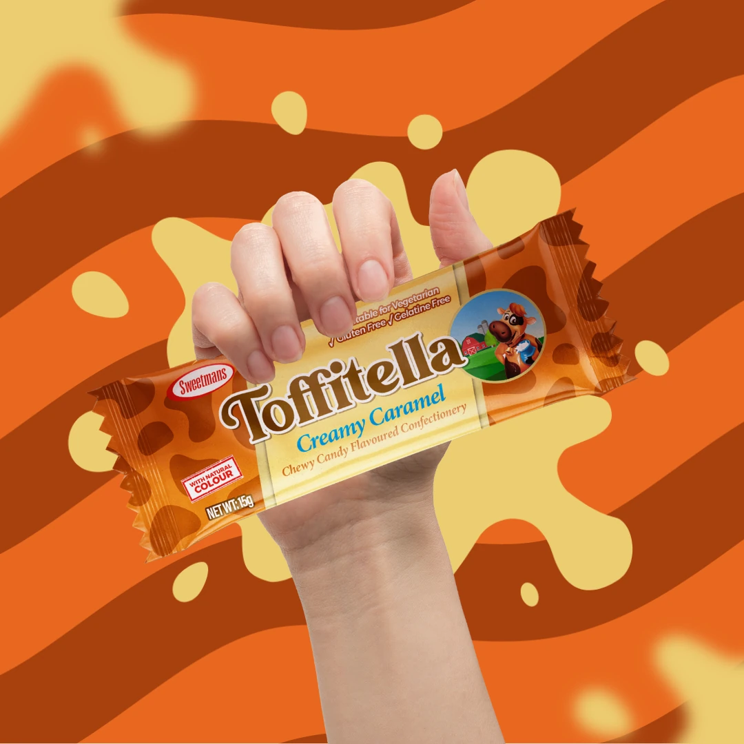 Hand holding a Sweetsman Toffitella Creamy Caramel candy bar against an orange and brown wavy background.