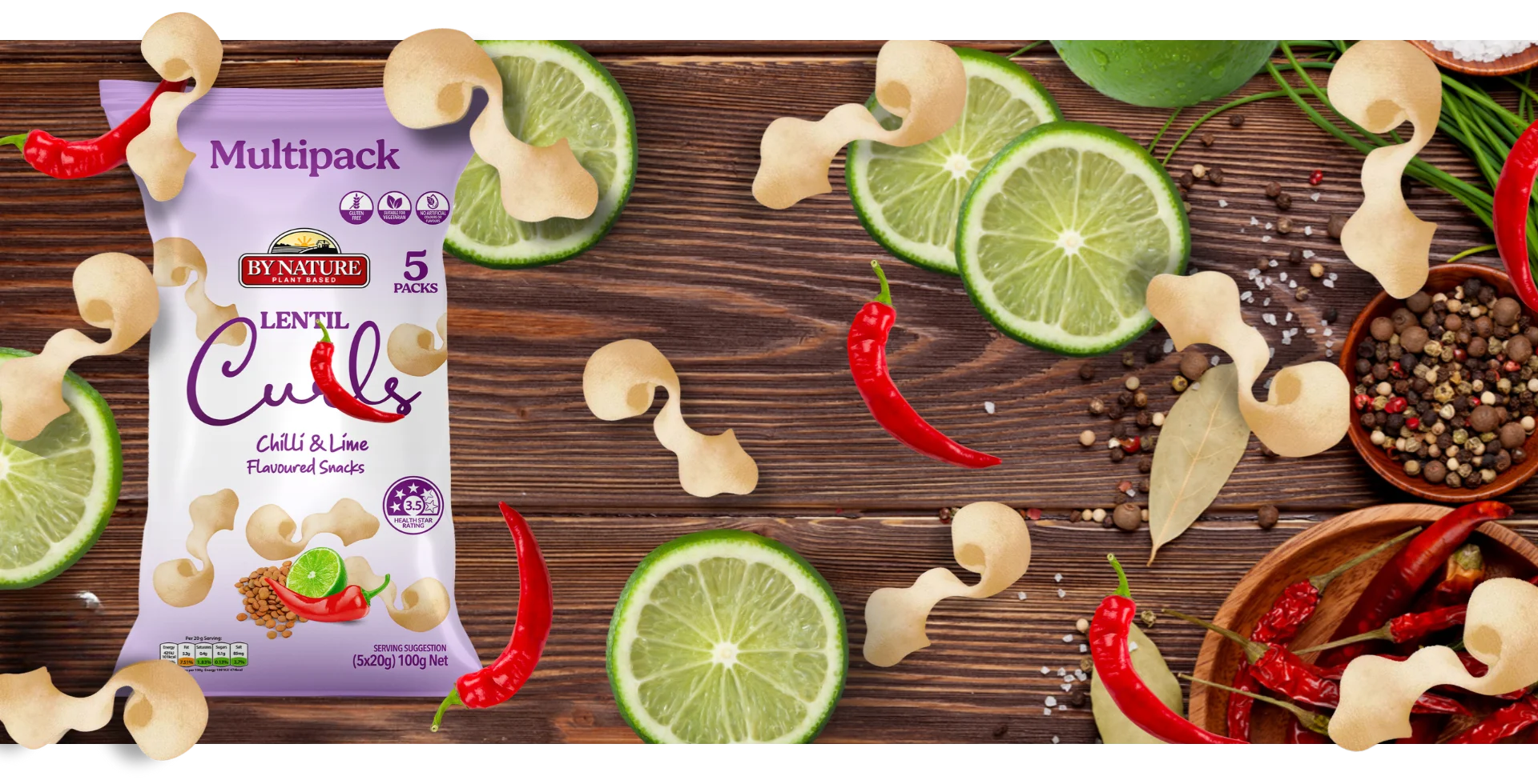 A stylised image of By Nature Lentil Curls Chilli & Lime packaging surrounded by chillies, lime slices, and seasoning on a wooden table.