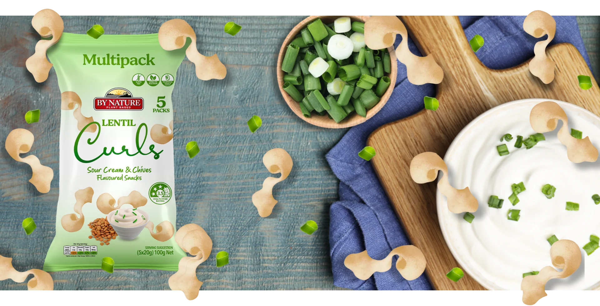 A stylised image of By Nature Lentil Curls Sour Cream & Chives packaging surrounded by chives and a bowl of sour cream on a wooden table.