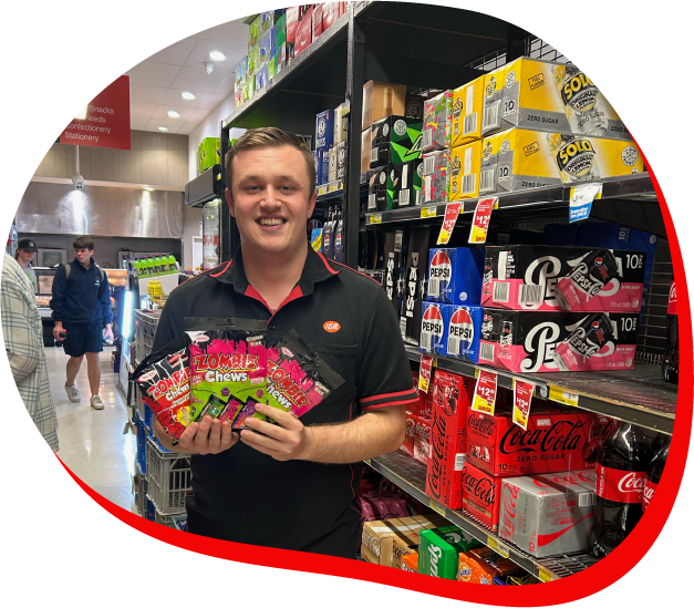 A man wearing an IGA uniform holding multiple packets of Zombie Chews in a supermarket aisle, with soda cans and other products in the background.