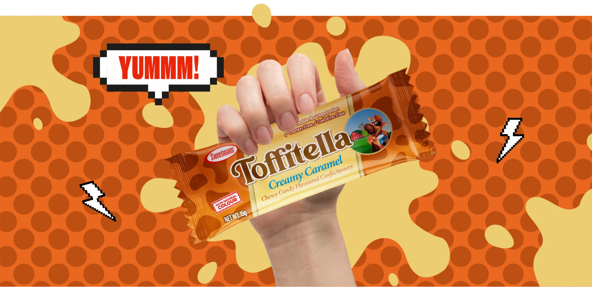 A hand holding a Toffitella creamy caramel chewy candy bar on a playful, dotted orange background with the word "Yummm!" in a pixelated speech bubble.