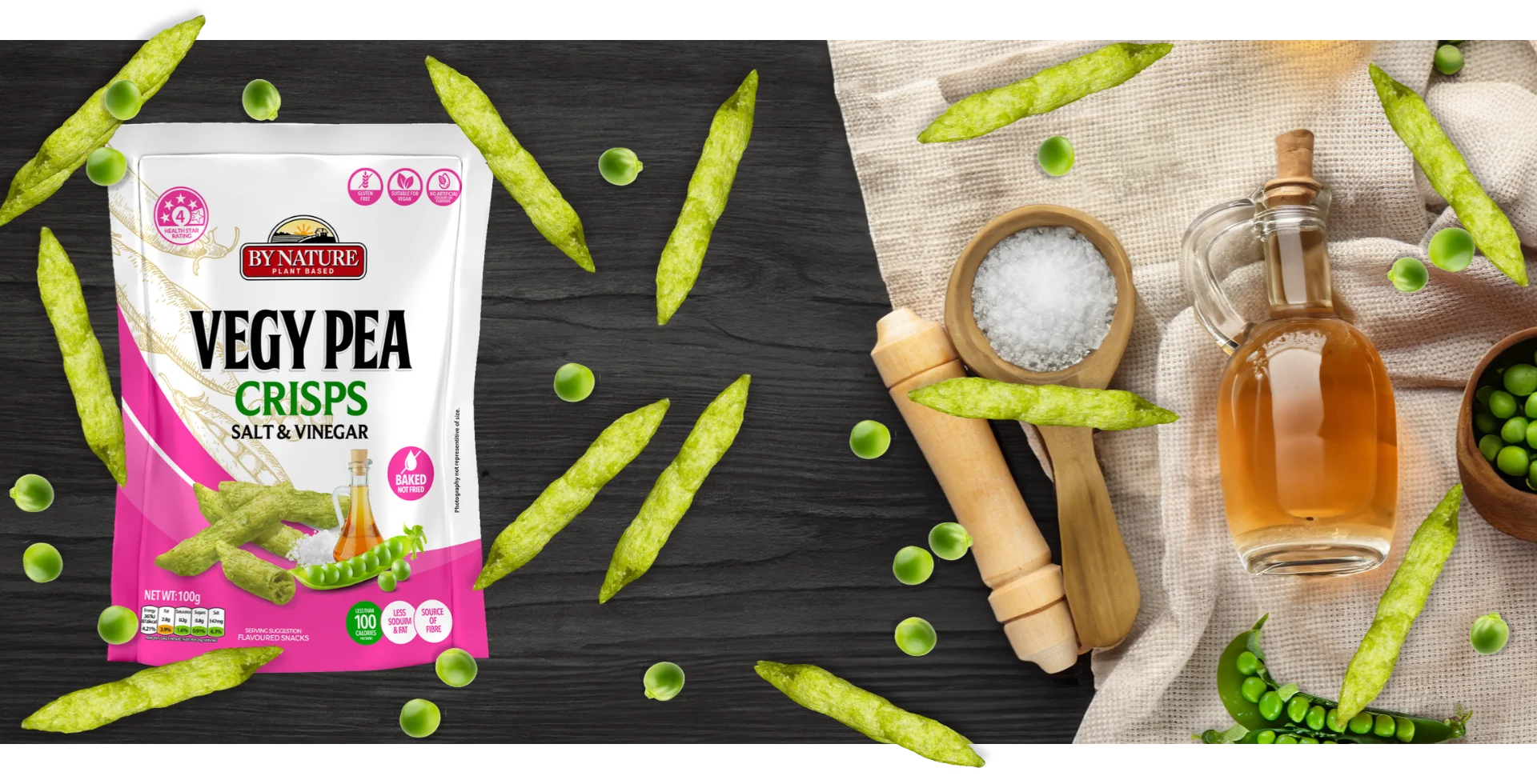 A stylised image of By Nature Vegy Pea Crisps Salt & Vinegar packaging surrounded by green peas, salt, and vinegar on a dark wooden table.