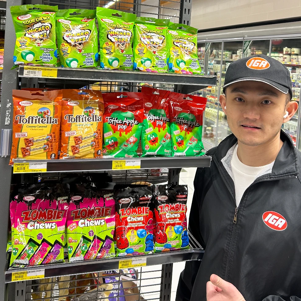 A man in an IGA uniform standing next to a store display shelf featuring Sweetmans products including Zombie Chews, Toffitella, and Toffee Apple Pop.